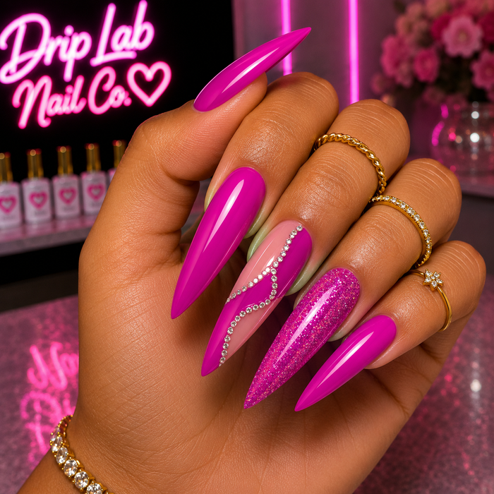 A close-up of a hand showcasing long, glossy stiletto-style press-on nails in vibrant shades of hot pink, magenta, and glittery pink, with one nail featuring a wavy silver rhinestone design. The background features a pink neon sign for 'Drip Lab Nail Co.