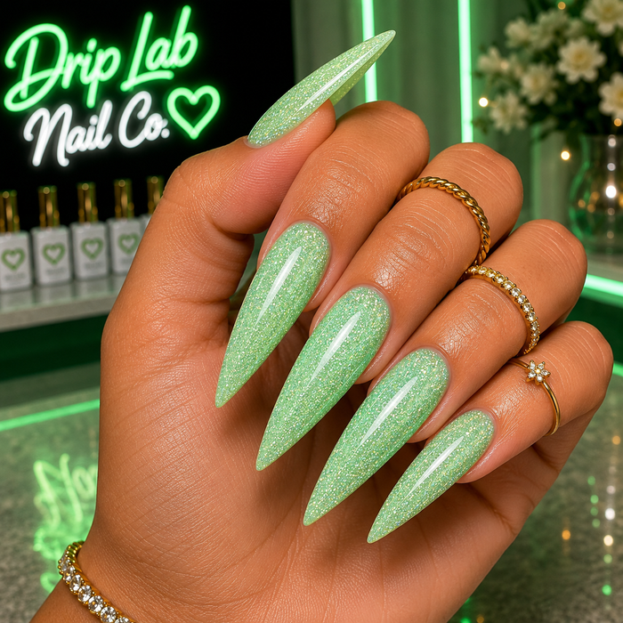 A close-up of a hand with long, stiletto-shaped nails painted in a light mint green glitter polish. The nails are shown on a person wearing gold rings and a bracelet, with a neon sign reading 'Drip Lab Nail Co.